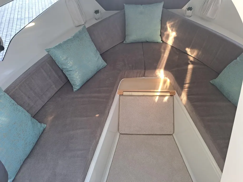Cosy interior of a boat featuring grey upholstery, teal cushions, and a small wooden table illuminated by sunlight.